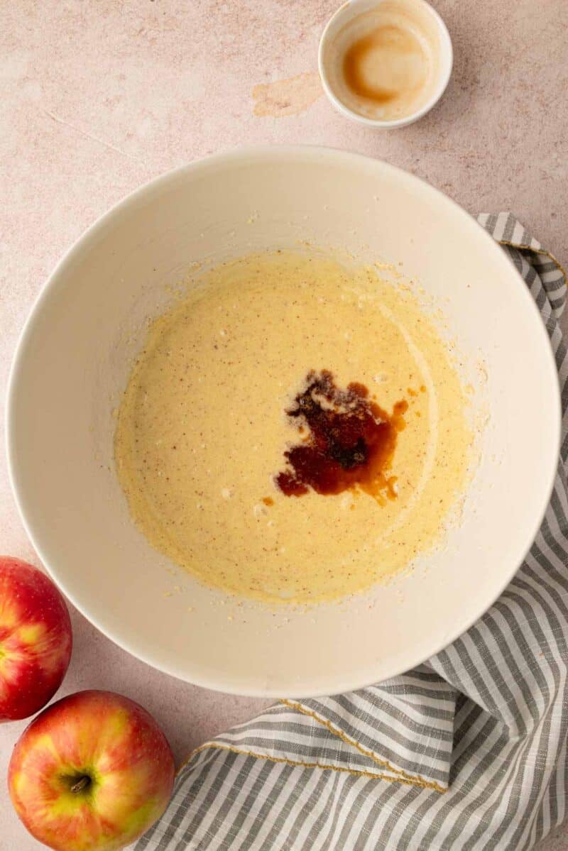 A mixing bowl with batter and a splash of vanilla extract, next to two apples, an empty small bowl, and a striped cloth on a light surface.