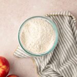 A glass bowl of flour sits on a gray and white striped cloth next to two red apples on a light countertop.