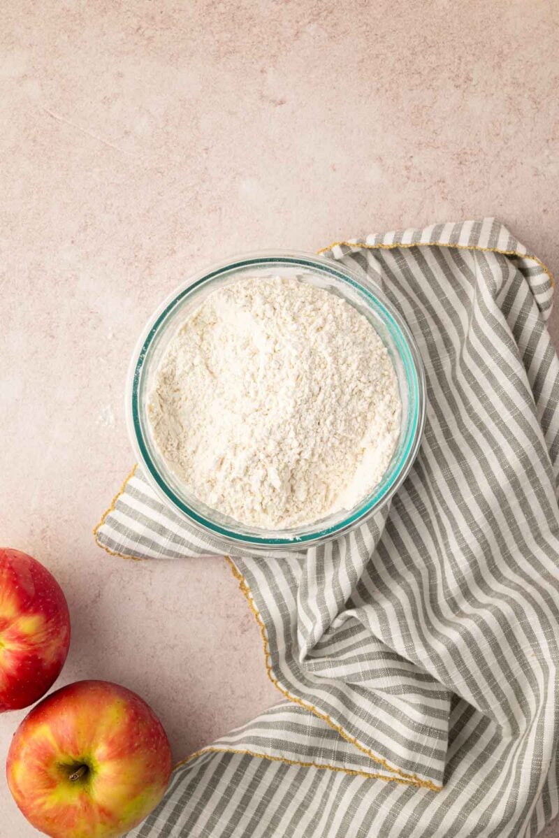 A glass bowl of flour sits on a gray and white striped cloth next to two red apples on a light countertop.