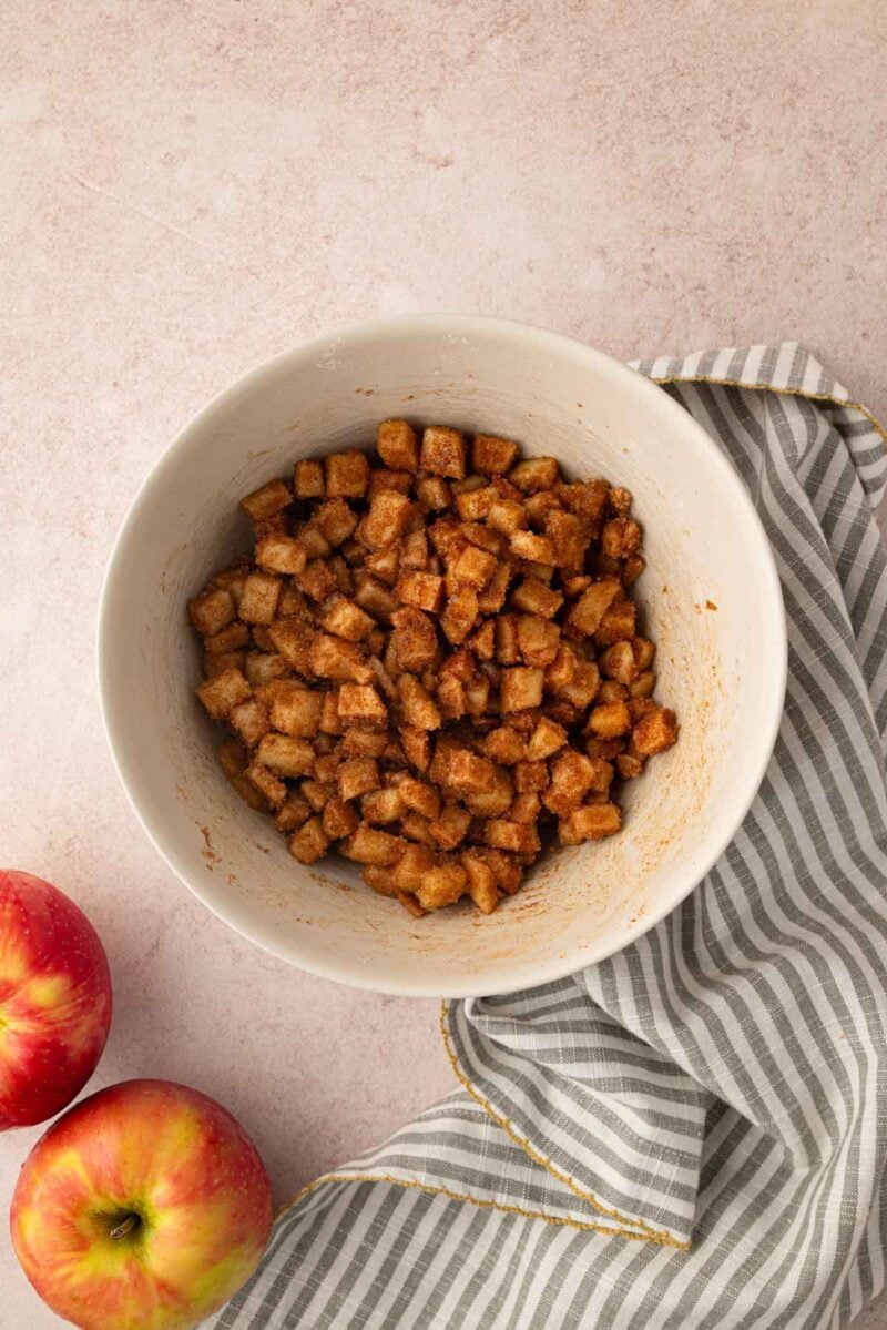 A bowl of cinnamon-coated diced apples sits on a light surface next to two whole apples and a gray-and-white striped cloth.