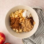 A bowl containing chopped apples, brown sugar, cinnamon, and flour sits on a striped cloth next to two whole apples on a light surface.
