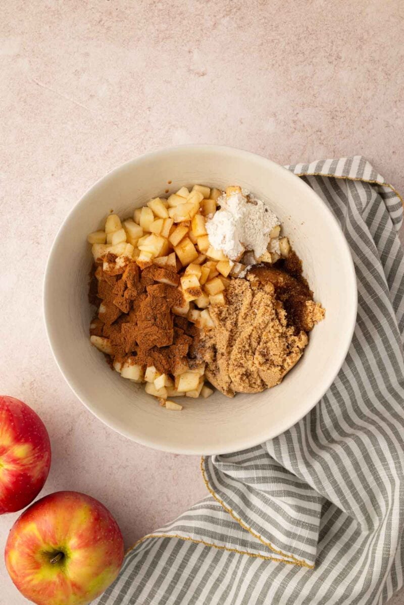A bowl containing chopped apples, brown sugar, cinnamon, and flour sits on a striped cloth next to two whole apples on a light surface.