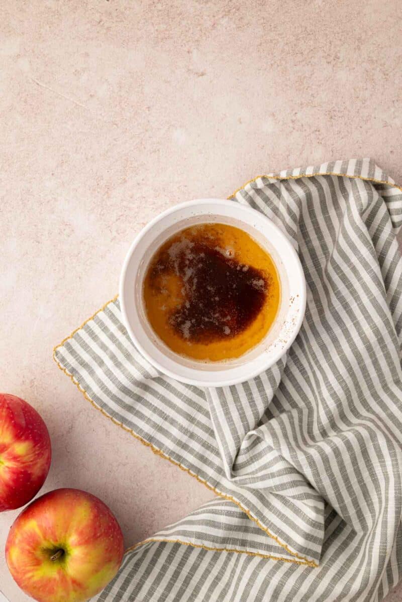 A bowl of melted butter on a striped cloth napkin, with two apples placed nearby on a light-colored surface.