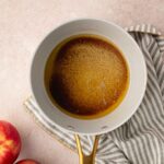 A saucepan with browned butter sits on a striped cloth next to two red apples on a light-colored surface.