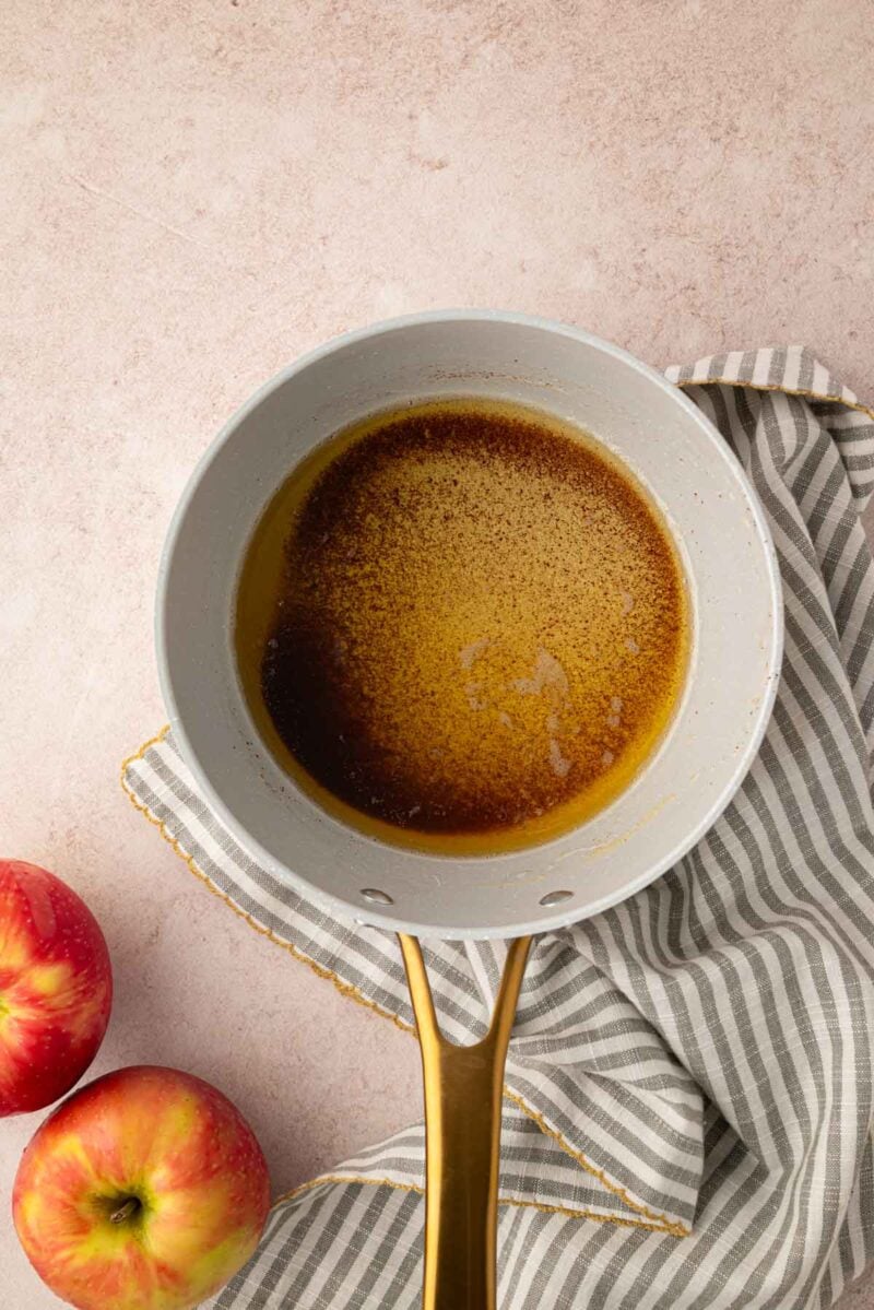 A saucepan with browned butter sits on a striped cloth next to two red apples on a light-colored surface.
