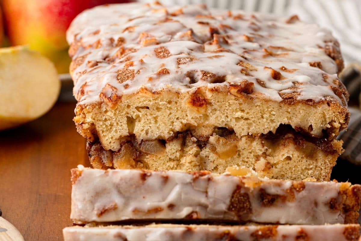 A sliced loaf of glazed apple bread sits on a wooden surface, revealing chunks of apple and a layer of cinnamon inside.