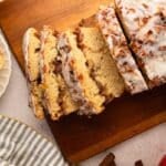 Sliced glazed apple cinnamon bread on a wooden cutting board, with a striped napkin, apple slices, and cinnamon sticks nearby.