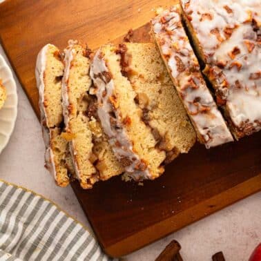 Sliced glazed apple cinnamon bread on a wooden cutting board, with a striped napkin, apple slices, and cinnamon sticks nearby.