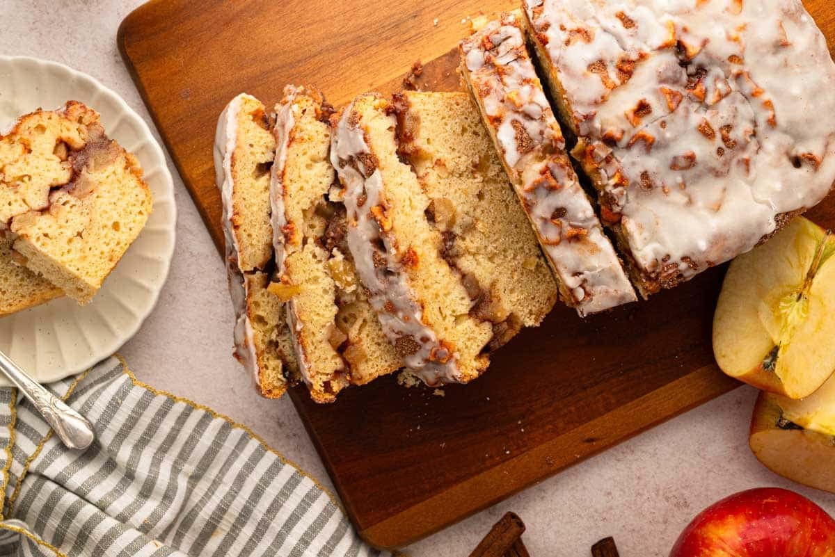 Sliced glazed apple cinnamon bread on a wooden cutting board, with a striped napkin, apple slices, and cinnamon sticks nearby.