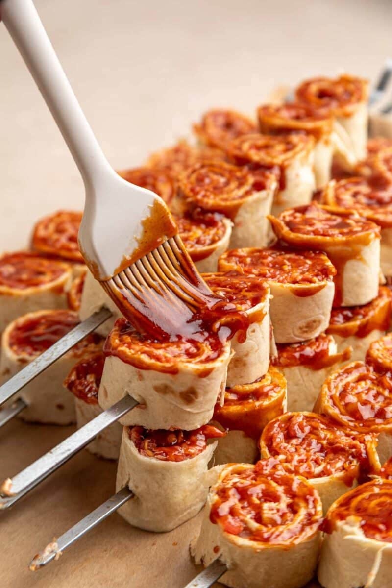 A brush applies sauce to rolled tortilla skewers, arranged closely together on a flat surface.