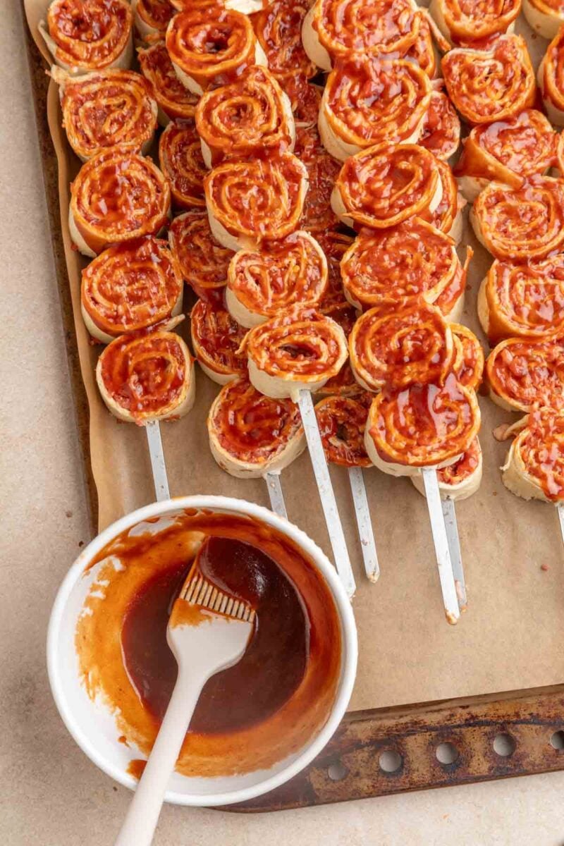 Unbaked bbq tortilla roll ups on skewers are arranged on a baking sheet lined with parchment paper, next to a bowl of pizza sauce and a pastry brush.