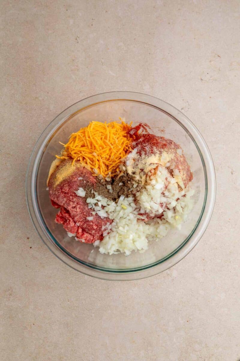 Glass bowl with ground beef, chopped onions, shredded cheddar cheese, seasonings, and breadcrumbs on a beige surface.