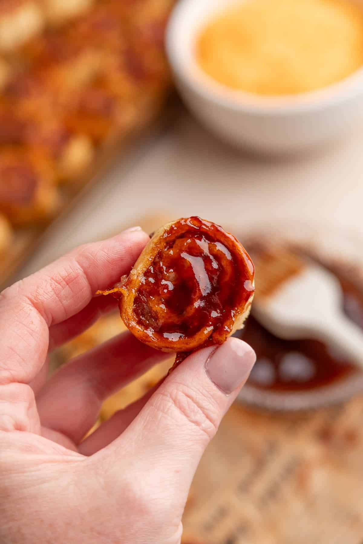 A hand holds a bite-sized bbq tortilla roll ups coated with barbecue sauce, with a bowl of sauce and other food items blurred in the background.