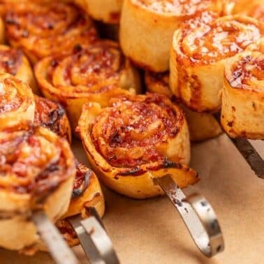 Close-up of bbq tortilla roll ups threaded onto metal skewers, showing melted cheese and tomato sauce on a brown surface.