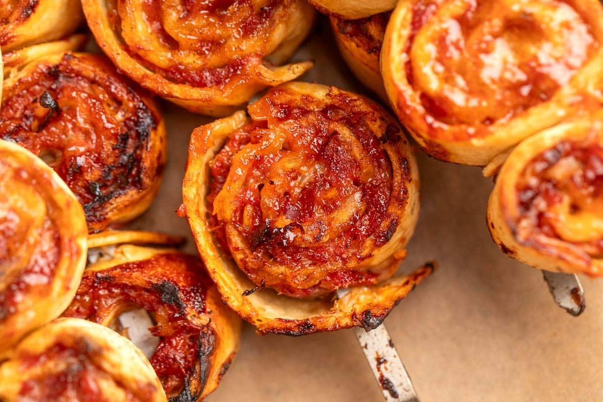 Close-up of several bbq tortilla roll ups with visible melted cheese and tomato sauce, arranged closely together on a brown surface.