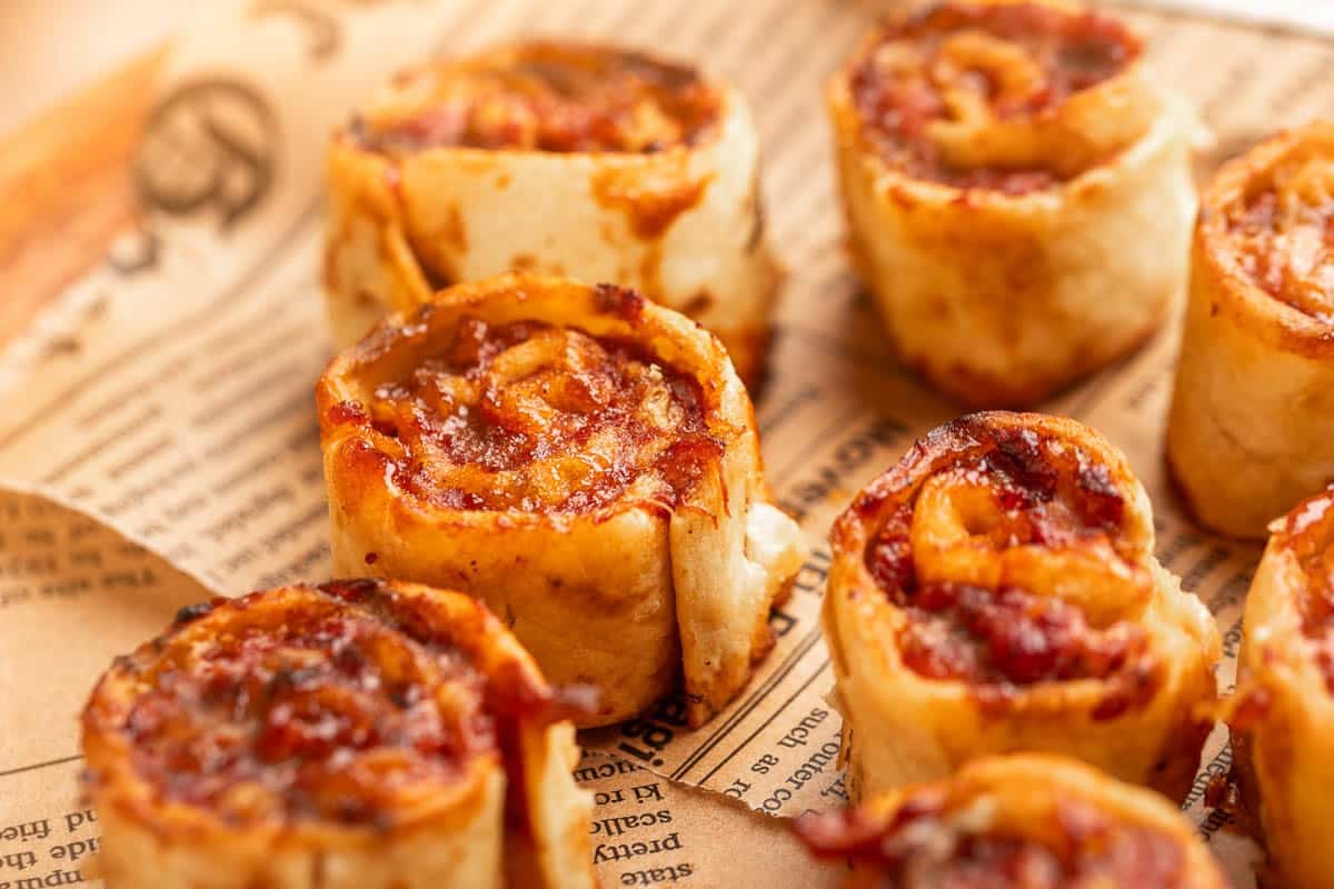 Close-up of several baked pinwheel snacks with a golden cheese and tomato filling, arranged on a sheet of printed parchment paper.