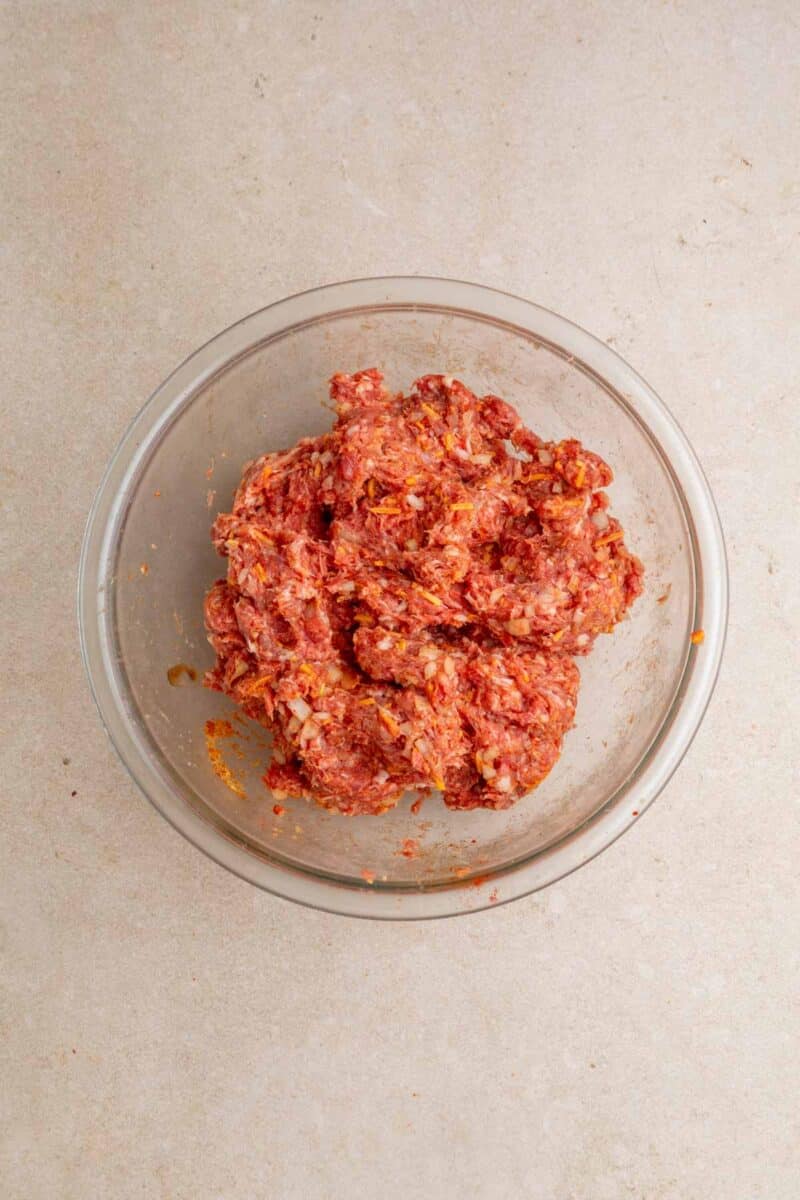 A glass bowl filled with raw ground meat mixed with chopped onions and seasonings sits on a light-colored countertop.