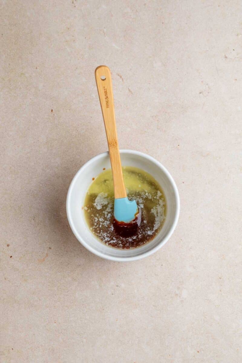 A small white bowl contains melted butter and honey, being stirred with a blue silicone spatula with a wooden handle on a light beige surface.
