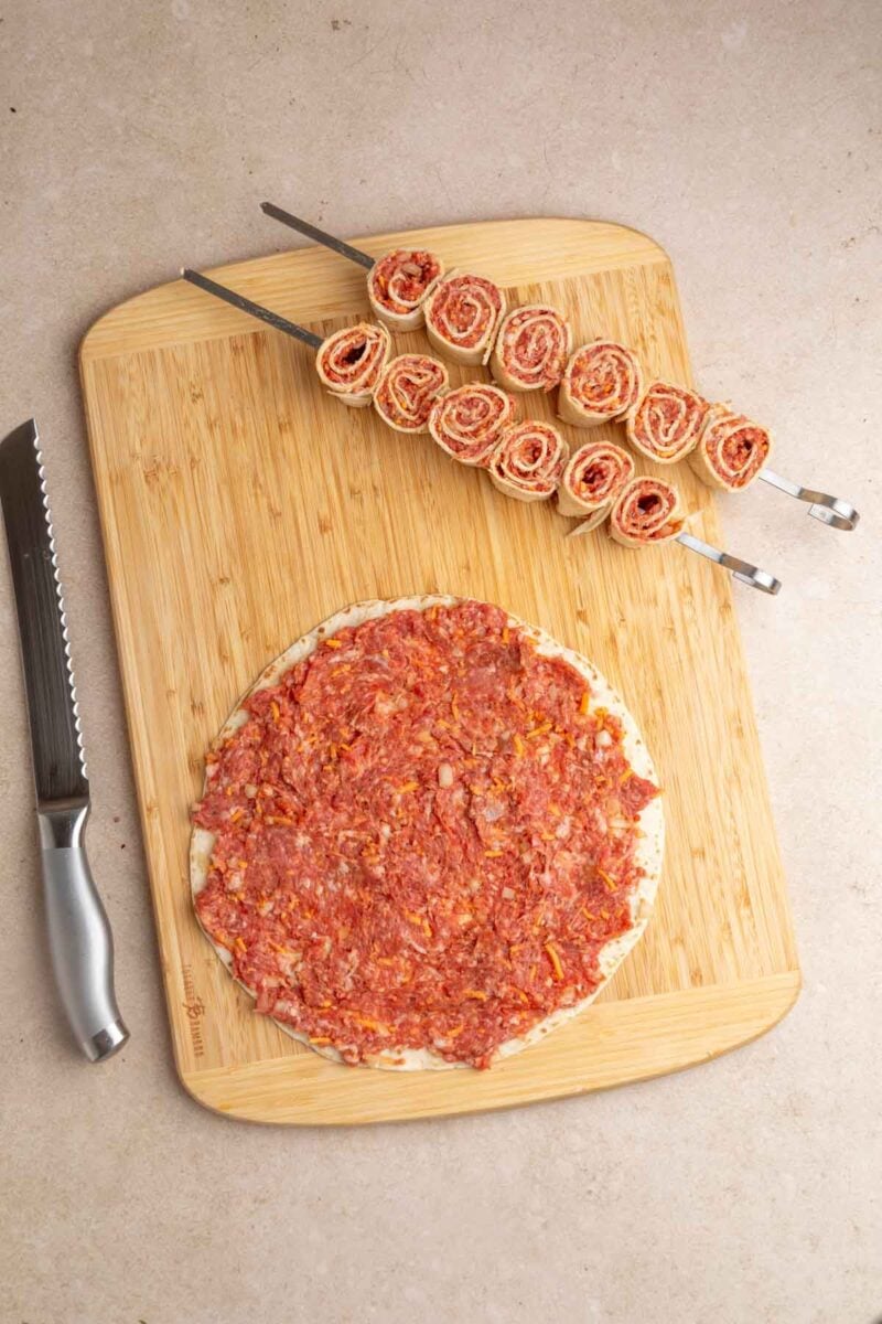 A round flatbread covered with a meat mixture is on a wooden cutting board next to two skewers of rolled pinwheel slices; a serrated knife is placed beside the board.
