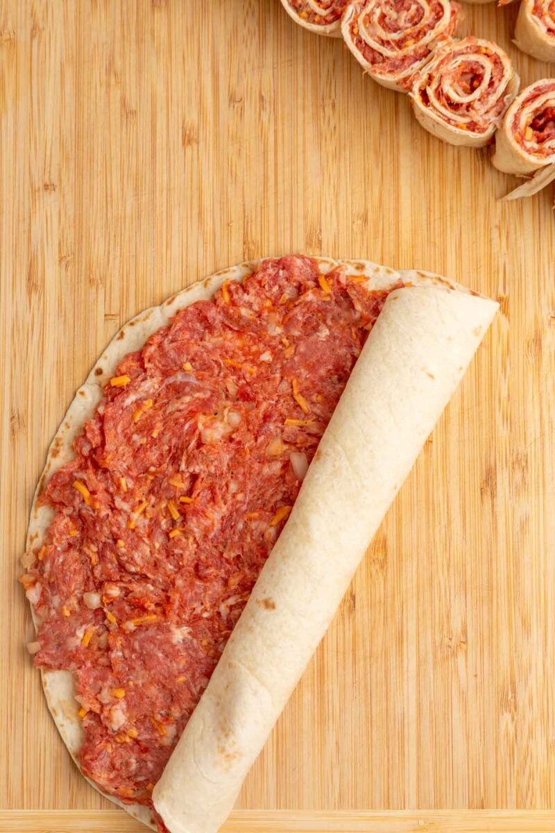 A tortilla spread with a ground beef mixture is being rolled up on a wooden board, with several completed pinwheels visible at the top.