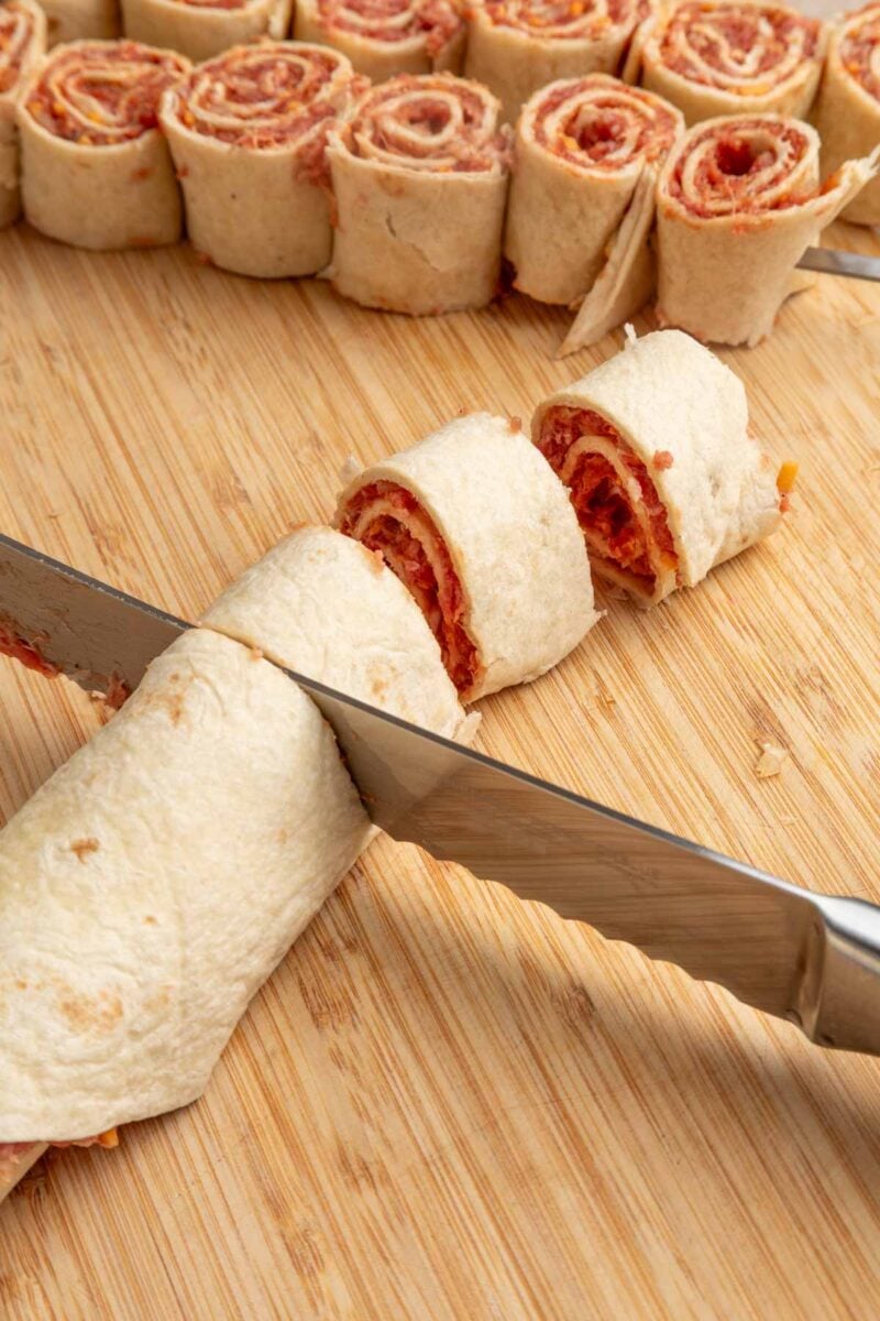 A knife slices a rolled tortilla with filling on a wooden cutting board; several rolled pinwheel pieces are visible in the background.