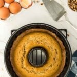 A baked bundt cake in a pan is on a cooling rack next to eggs, butter, chopped pecans, and a cake server on a marble countertop.