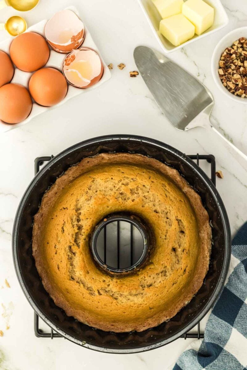 A baked bundt cake in a pan is on a cooling rack next to eggs, butter, chopped pecans, and a cake server on a marble countertop.
