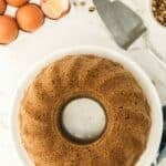 A whole bundt cake on a white plate, surrounded by eggs, butter, chopped nuts, and a cake server on a marble countertop.
