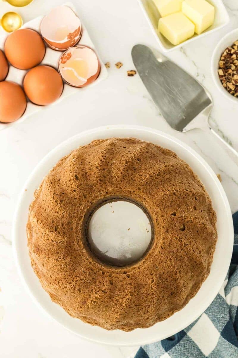 A whole bundt cake on a white plate, surrounded by eggs, butter, chopped nuts, and a cake server on a marble countertop.
