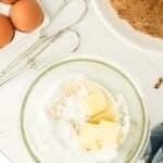 A glass bowl with butter and powdered sugar, eggs, a hand mixer, chopped pecans, and a bundt cake on a white surface.