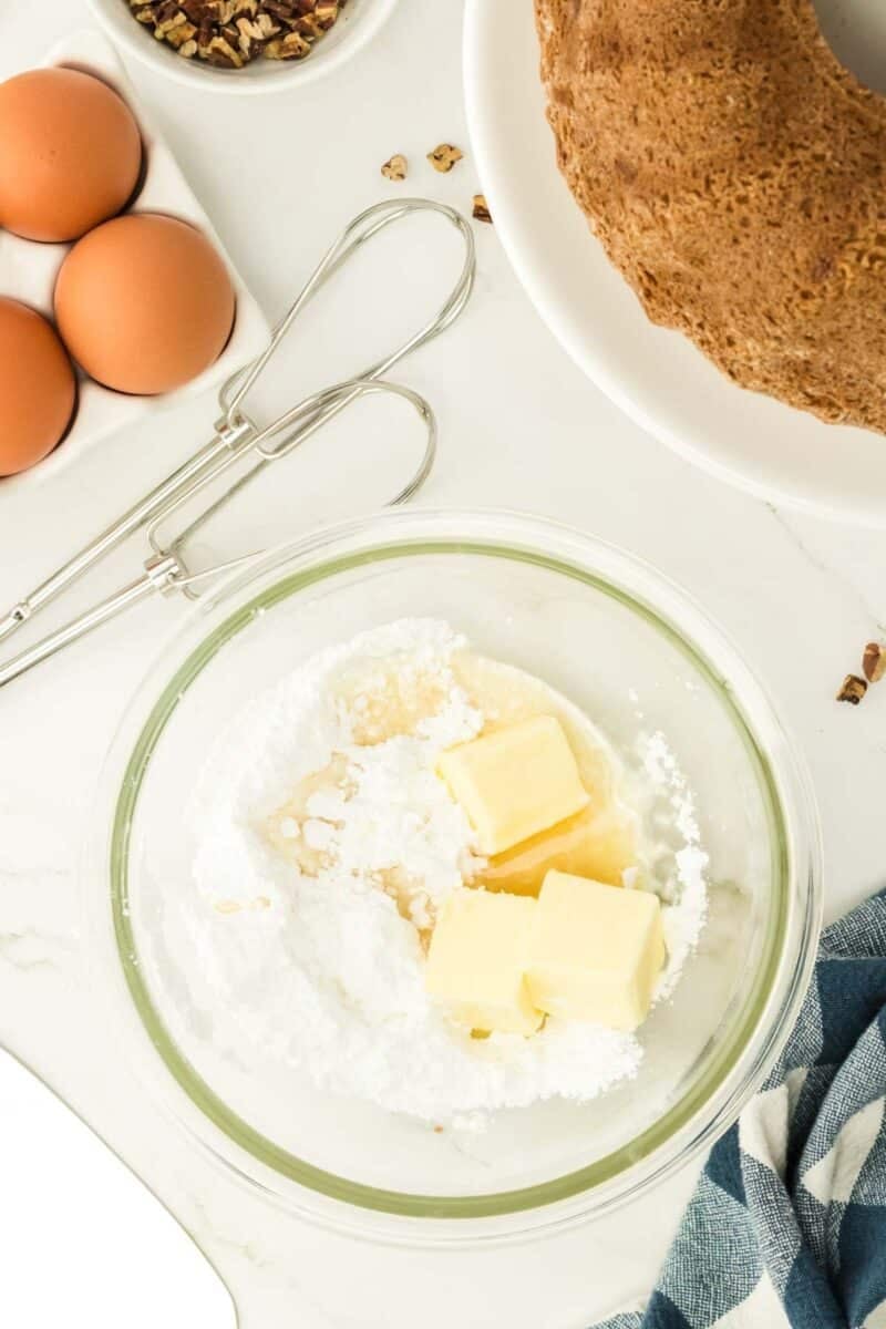 A glass bowl with butter and powdered sugar, eggs, a hand mixer, chopped pecans, and a bundt cake on a white surface.