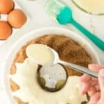 A hand spoons white icing over a bundt cake on a white plate, with eggs, chopped nuts, and baking tools nearby on a white countertop.