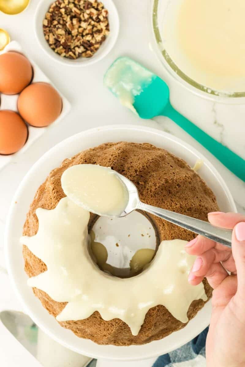 A hand spoons white icing over a bundt cake on a white plate, with eggs, chopped nuts, and baking tools nearby on a white countertop.