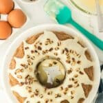 A bundt cake with white icing and chopped nuts on top sits on a plate. Eggs, a bowl of nuts, a green spatula, and a bowl of icing are nearby on a marble surface.