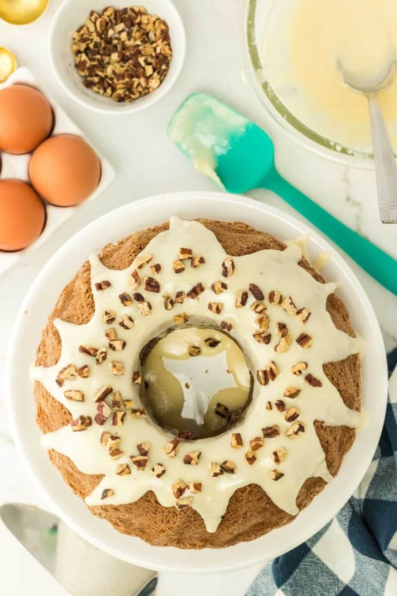 A bundt cake with white icing and chopped nuts on top sits on a plate. Eggs, a bowl of nuts, a green spatula, and a bowl of icing are nearby on a marble surface.