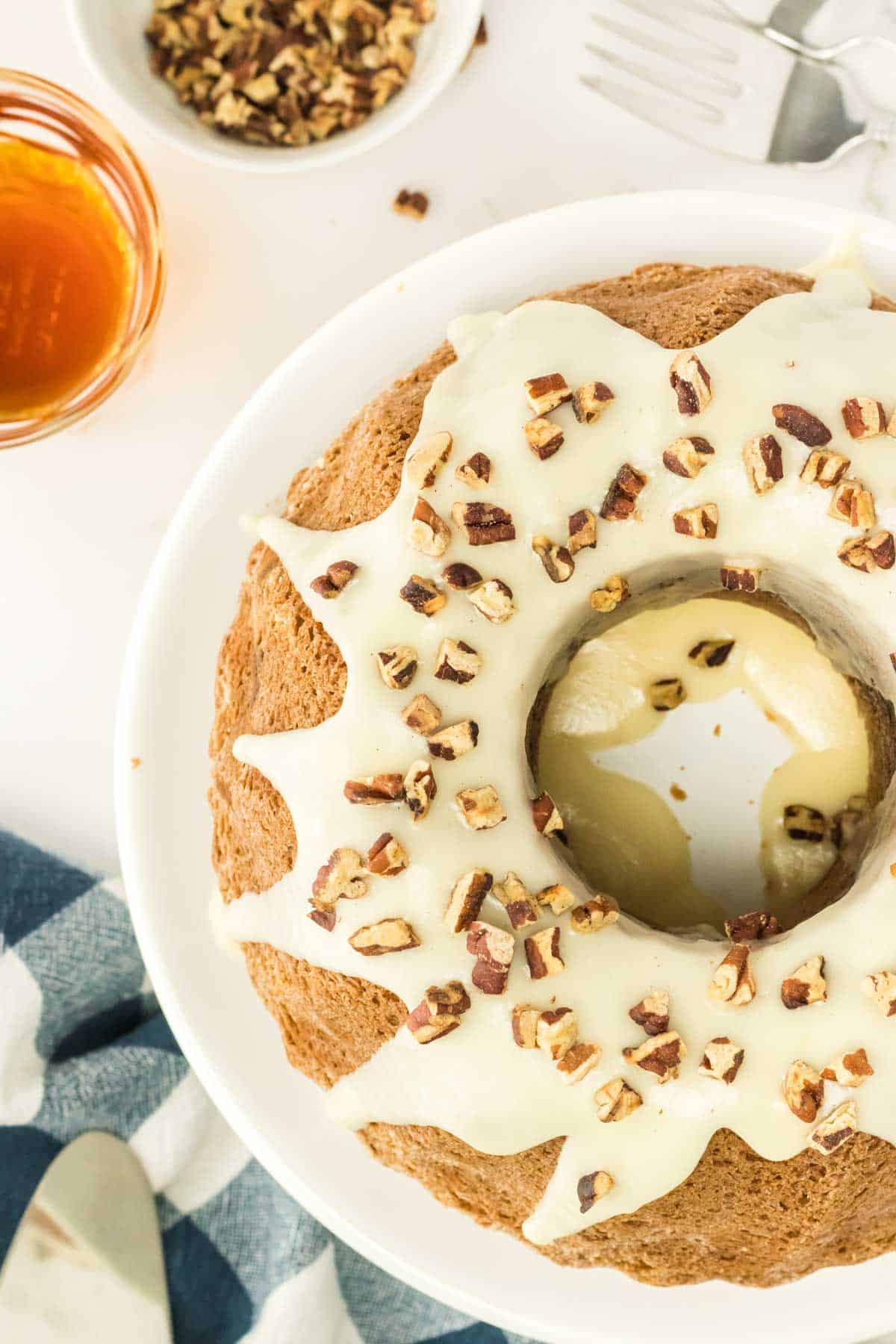 Bundt cake with white icing and chopped pecans on a white plate, shown with a bowl of pecans, a glass of honey, and utensils nearby.