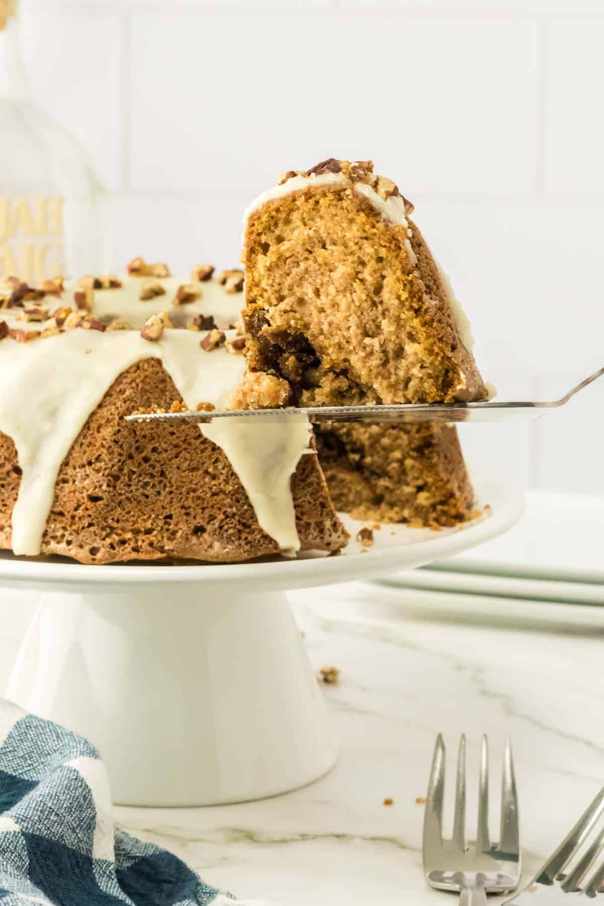 A slice of bundt cake with icing and chopped nuts is being lifted from the cake, which sits on a white cake stand. Forks and a cloth napkin are on the marble counter nearby.