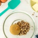 Glass bowl with brown sugar and chopped pecans on a marble surface, surrounded by eggs, butter, a teal spatula, a striped cloth, and gold measuring spoons.