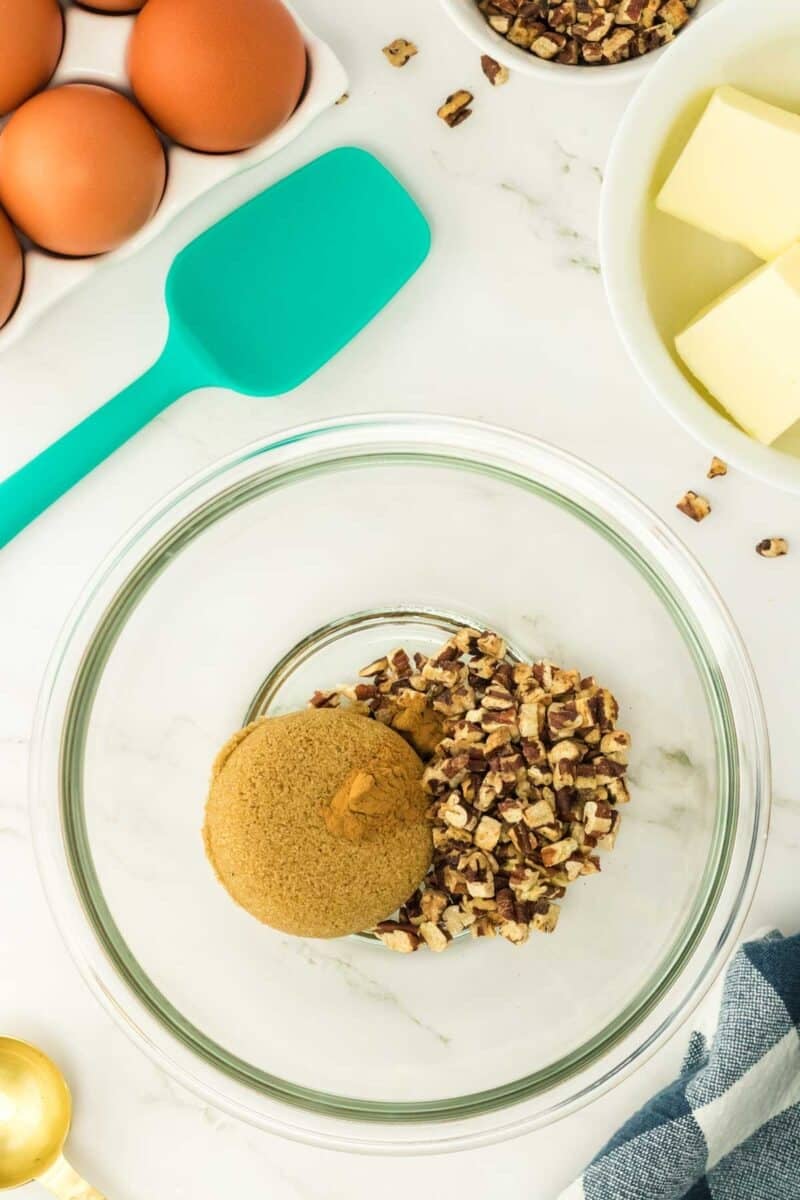 Glass bowl with brown sugar and chopped pecans on a marble surface, surrounded by eggs, butter, a teal spatula, a striped cloth, and gold measuring spoons.
