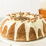 Bundt cake with white icing and chopped nuts on top, displayed on a white cake stand. A bottle labeled “Elijah Craig” is visible in the background.