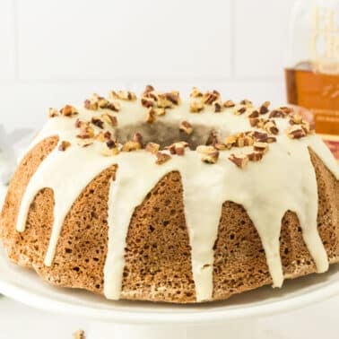 Bundt cake with white icing and chopped nuts on top, displayed on a white cake stand. A bottle labeled “Elijah Craig” is visible in the background.