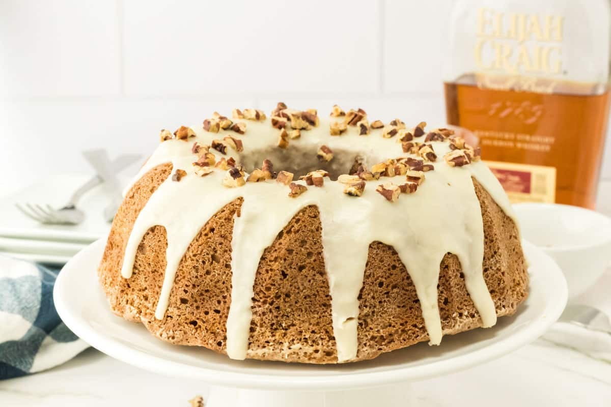 Bundt cake with white icing and chopped nuts on top, displayed on a white cake stand. A bottle labeled “Elijah Craig” is visible in the background.
