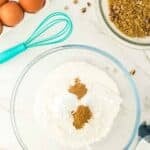 A glass bowl with flour and spices, a whisk, eggs, chopped pecans, brown sugar mixture, measuring spoons, and a blue checked cloth on a white surface.
