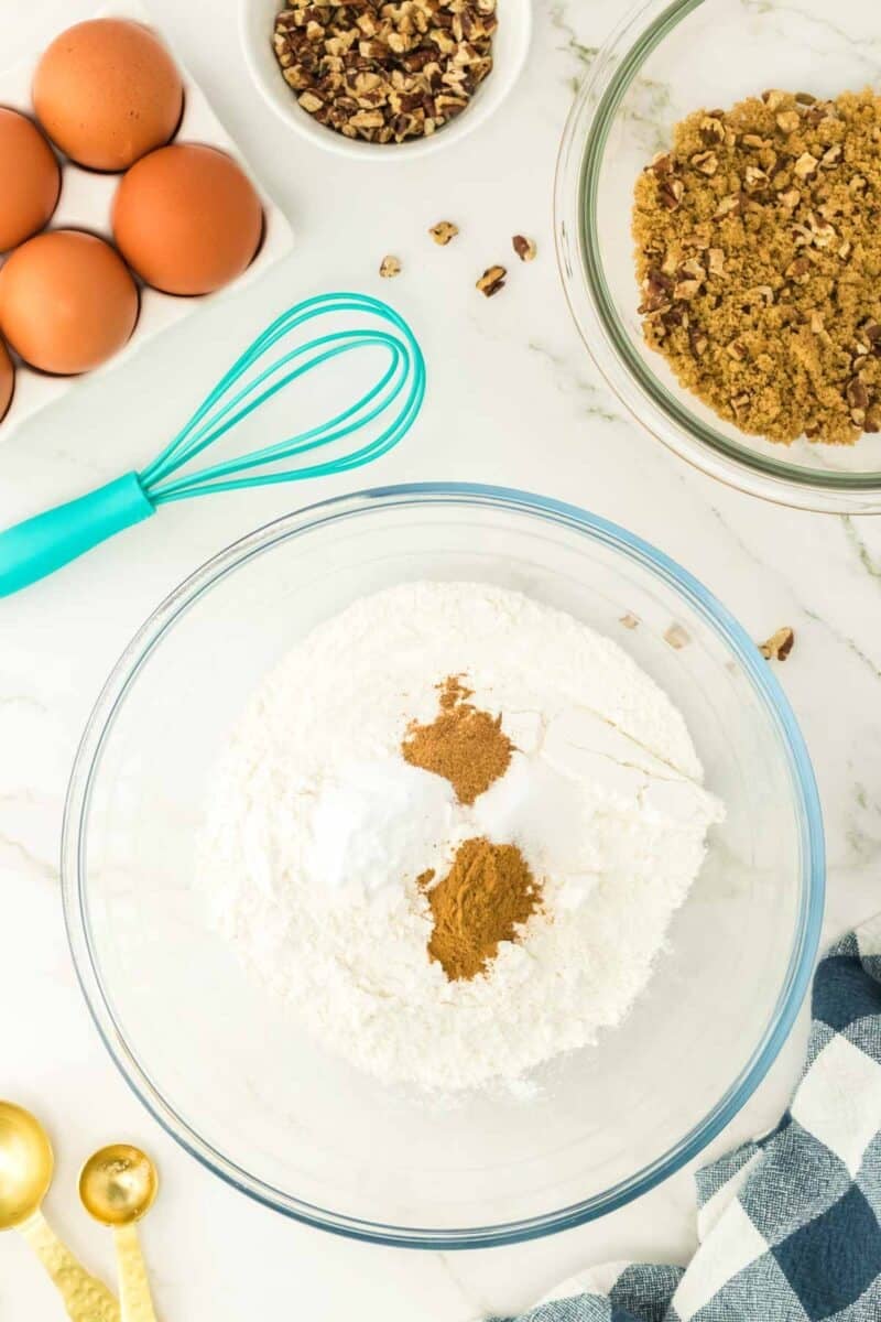 A glass bowl with flour and spices, a whisk, eggs, chopped pecans, brown sugar mixture, measuring spoons, and a blue checked cloth on a white surface.