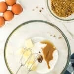 A glass bowl with butter, sugar, and vanilla, an electric mixer, whole eggs, chopped pecans, and brown sugar on a marble countertop.