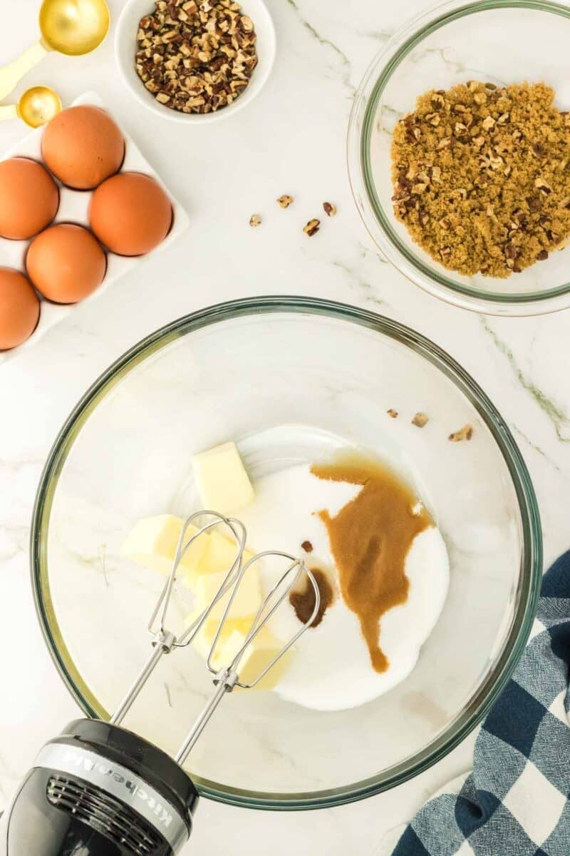 A glass bowl with butter, sugar, and vanilla, an electric mixer, whole eggs, chopped pecans, and brown sugar on a marble countertop.