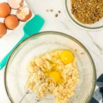 Electric mixer in a glass bowl with cookie dough and two eggs, surrounded by eggshells, chopped nuts, and mixing tools on a marble countertop.