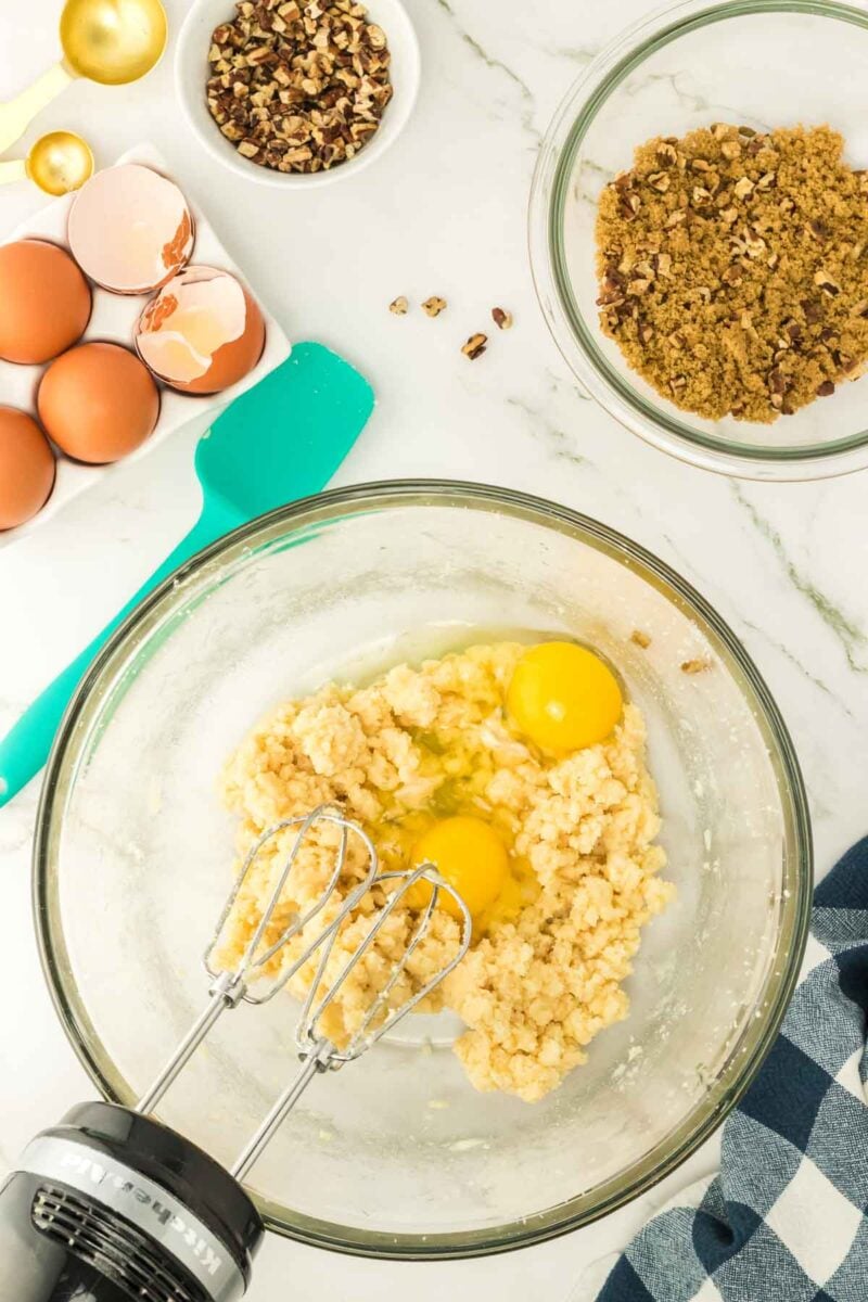 Electric mixer in a glass bowl with cookie dough and two eggs, surrounded by eggshells, chopped nuts, and mixing tools on a marble countertop.