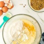 Glass mixing bowl with flour, eggs, and butter being mixed by a hand mixer, surrounded by eggs, chopped pecans, and a bowl of brown sugar on a marble countertop.