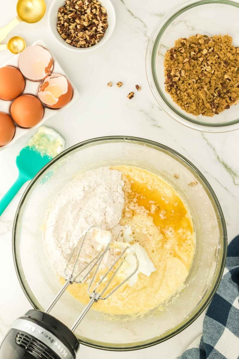 Glass mixing bowl with flour, eggs, and butter being mixed by a hand mixer, surrounded by eggs, chopped pecans, and a bowl of brown sugar on a marble countertop.